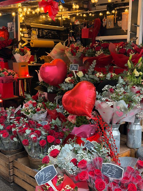 A vibrant floral display at a flower shop showcasing an array of fresh red roses, white baby's breath, and greenery arranged in bouquets and wrapping paper. Heart-shaped foil balloons in red are featu