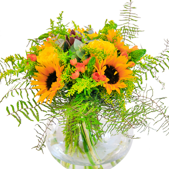This exuberant bouquet evokes the brightness of a summer's day in Anerley, as if the sun had settled gently amongst a lavish collection of blooms. Striking sunflowers, with their golden rays and chocolate centres, serve as the heart of the arrangement, their large faces directing light and warmth to every corner. Around them, soft yellow garden roses unfurl with precision, their tight, velvet petals emitting a subtle fragrance that recalls walks through local gardens in bloom. Sprigs of glistening red Hypericum berries peek from within, their pops of colour infusing the composition with cheerful energy and playful charm. The bouquet is woven through with rich greenery: feathery ferns trail and arch with fluid movement, slender spikes and air-light fillers reminiscent of wildflowers found in the open spaces of Anerley. Silvery eucalyptus leaves, cool against the bright blooms, lend an understated touch of sophistication. Gathered in a clear, round glass vase, the bouquet's stems form a vivid tangle below the waterline, grounding the design and highlighting the freshness that Anerley expects from its neighbourhood florists. This artful arrangement brims with seasonal character, perfect for marking joyful milestones or adding a splash of happiness to everyday moments. Its thoughtful craftmanship, inspired by local parks and gardens, makes it a favourite for those seeking to brighten a home or heart in Anerley.